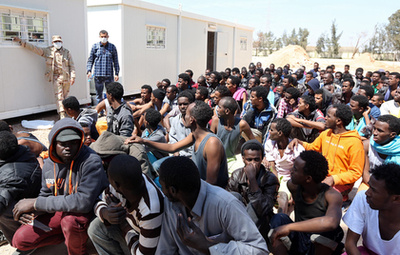 Egy olasz tiszt és az ideiglenes táborukat elhagyó menekültek Lampedusa repülőterén. Mivel az apró sziget kapacitásai végesek, a befogadott menekülteket innen az olasz szárazföldre szállítják.