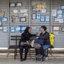 Két nő várakozik egy buszmegállóban, ahol gyászjelentéseket ragasztottak ki mögöttük Kriveljben, Szerbiában 2024. április 3-án