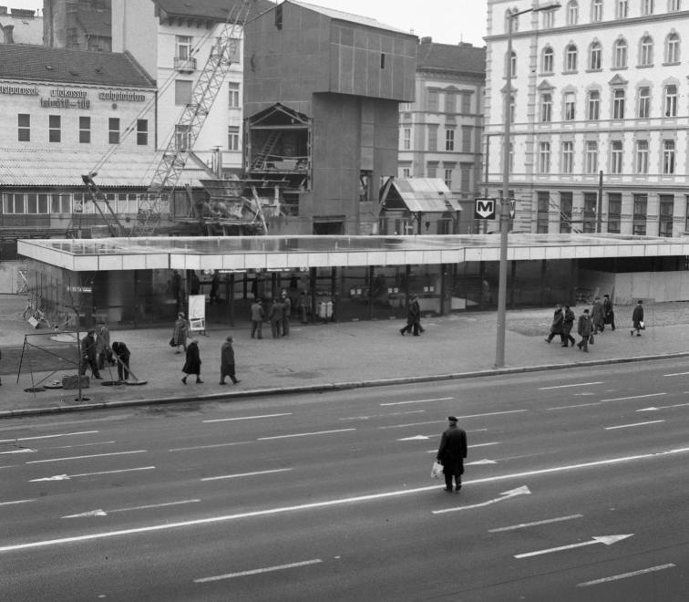 Még nem hívták Podmaniczky térnek az Arany János utcai metrómegálló felszíni területét, amikor 1982-ben ez a fotó elkészült. Négy munkás vizsgálja az újonnan megnyílt állomáslejáró elé kihelyezett utasforgalmi táblát, miközben egy sapkás úr magyaráz nekik.